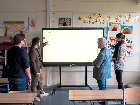 Vier Personen stehen in einem Klassenzimmer vor einem erleuchteten Smartboard und schauen darauf