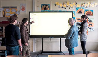 Vier Personen stehen in einem Klassenzimmer vor einem erleuchteten Smartboard und schauen darauf